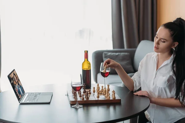 Junge Frau spielt Schach während Videotelefonie mit Freund in der Nähe von Rotwein auf dem Tisch — Stockfoto