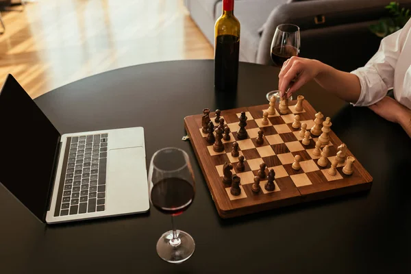 Vista recortada de la mujer jugando al ajedrez cerca de la computadora portátil con pantalla blabk y vino tinto en la mesa - foto de stock