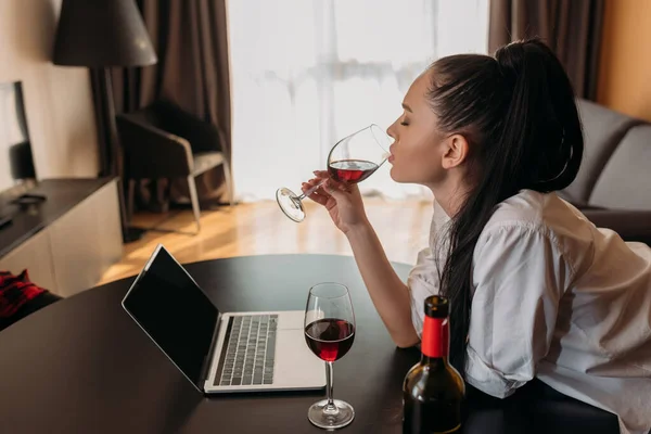 Vue latérale de la jeune femme buvant du vin rouge près d'un ordinateur portable avec écran blanc — Photo de stock
