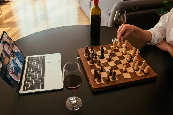 Vista recortada de la mujer jugando al ajedrez durante una videollamada con su novio cerca de vino tinto en la mesa - foto de stock