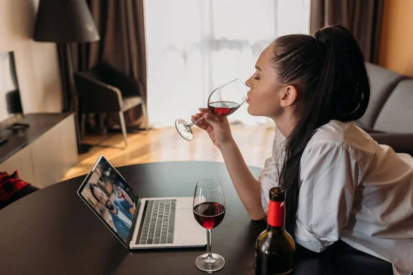 Vue latérale de la jeune femme buvant du vin rouge pendant le chat vidéo avec son petit ami — Photo de stock