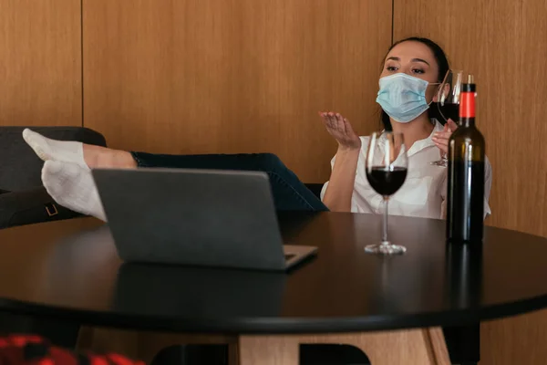 Jeune femme en masque médical envoyer baiser d'air pendant le chat vidéo près de la bouteille et des verres de vin rouge — Photo de stock