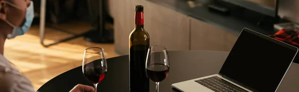 Cropped view of young woman in medical mask near laptop with blank screen, glasses ad bottle of red wine, panoramic shot — Stock Photo