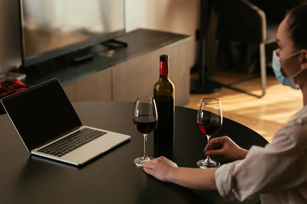 Foco seletivo da jovem mulher em máscara médica perto de laptop com tela em branco, óculos e garrafa de vinho tinto — Fotografia de Stock