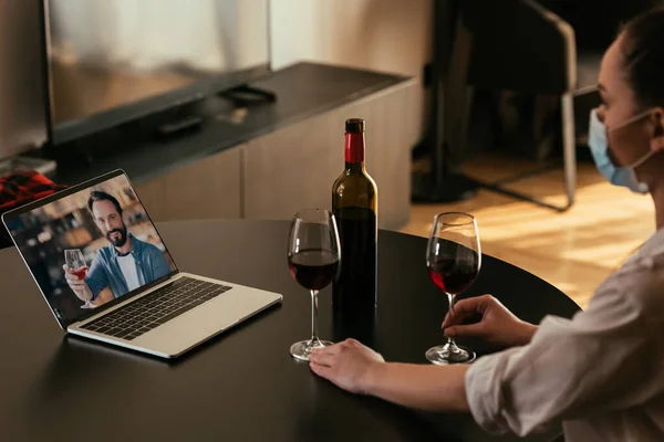 Messa a fuoco selettiva di giovane donna in maschera medica avendo video chat con fidanzato vicino bicchieri e bottiglia di vino rosso — Foto stock