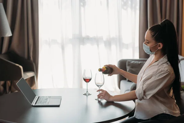 Vista lateral da jovem mulher em máscara protetora derramando vinho em vidro perto do laptop com tela em branco — Fotografia de Stock