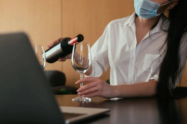 Vista ritagliata od donna in maschera medica versando vino in vetro vicino laptop — Foto stock