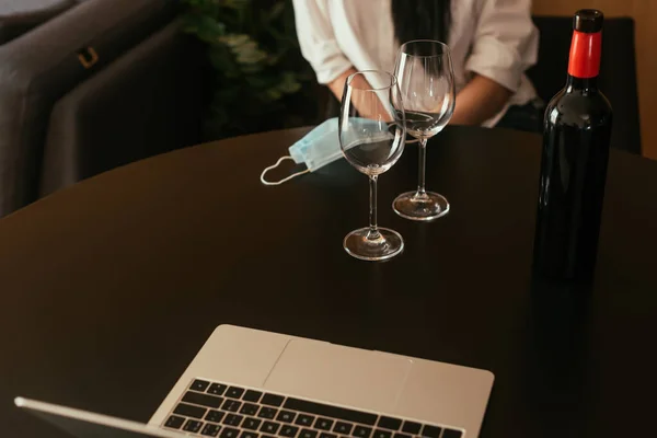 Обрезанный вид женщины, сидящей за столом рядом бокалы вина, бутылка, медицинская маска и ноутбук — стоковое фото