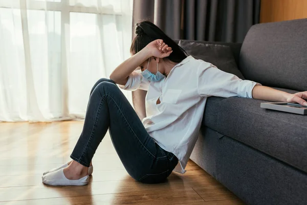 Grieving young woman in medical mask obscuring face with hand while sitting on floor and touching photo frame — Stock Photo