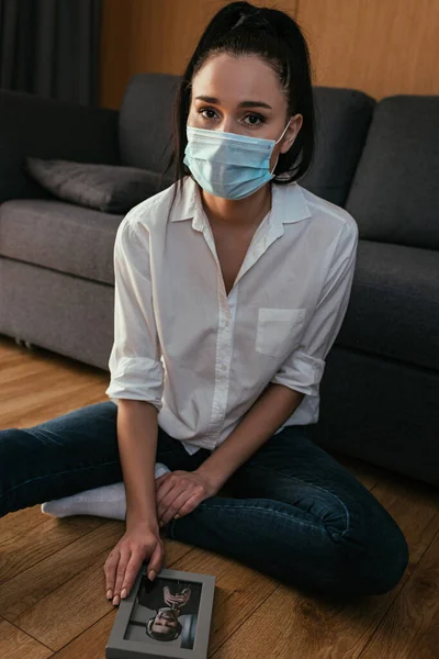 Stressed girl in a medical mask looking at camera while sitting on floor with photo of young man — Stock Photo