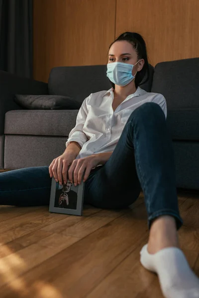 Selective focused of depressed woman in protective mask sitting on floor with photo of young man — Stock Photo