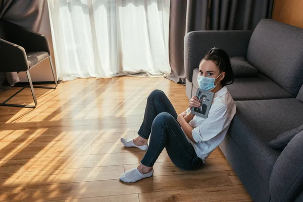 Fille déprimée dans un masque médical assis sur le sol avec photo de petit ami et regardant la caméra — Photo de stock