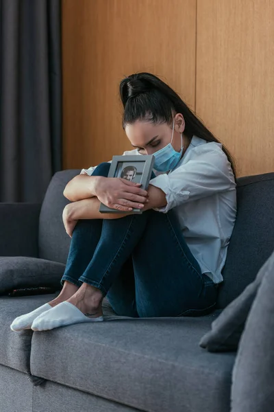 Mujer joven en duelo máscara médica sosteniendo foto de novio con mientras está sentado en el sofá con los ojos cerrados - foto de stock