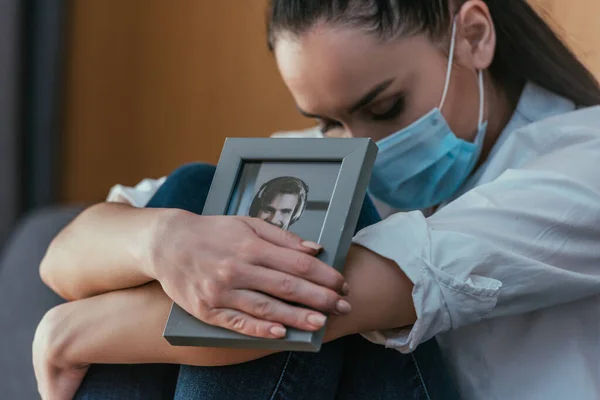 Afligida joven en máscara médica sosteniendo foto de novio con mientras está sentado con los ojos cerrados - foto de stock