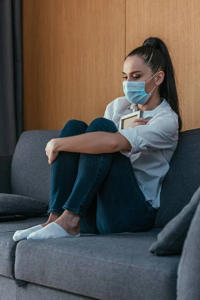 Jeune femme déprimée dans le masque médical tenant cadre photo près de la poitrine tout en étant assis sur le canapé — Photo de stock