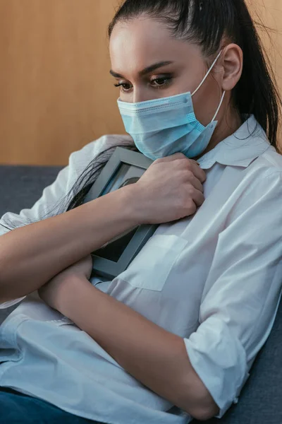 Mujer joven estresada en la máscara médica de duelo mientras sostiene el marco de fotos cerca del pecho - foto de stock