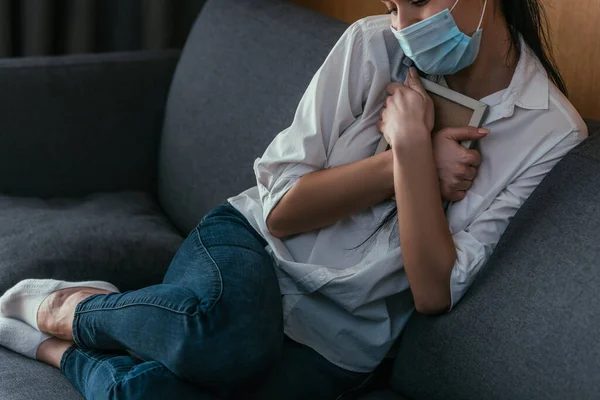 Обрізаний вид на депресивну молоду жінку в медичній масці, яка сумує, тримаючи фоторамку біля грудей, сидячи на дивані — стокове фото