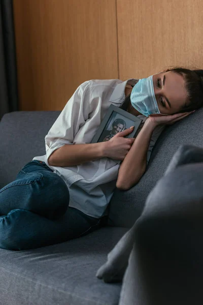 Mujer joven deprimida en máscara médica sosteniendo la foto del novio cerca del pecho mientras está sentado en el sofá - foto de stock