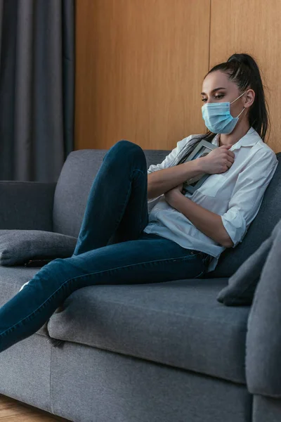 Triste jeune femme dans un masque de protection tenant cadre photo près de la poitrine tout en étant assis sur le canapé — Photo de stock