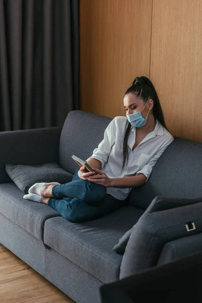 Depressed young woman in medical mask looking at photo frame while sitting on sofa — Stock Photo