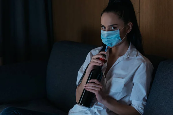 Депрессивная женщина в медицинской маске держит бутылку вина и смотрит в камеру — стоковое фото