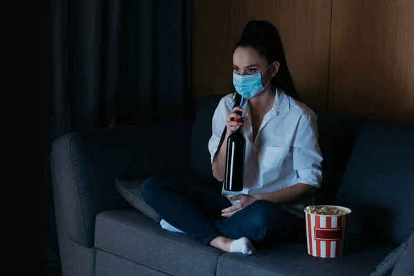 Подавленная женщина в медицинской маске смотрит телевизор, сидя на диване возле ведра попкорна и держа бутылку вина — стоковое фото