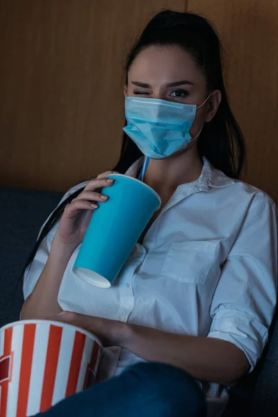 Молода жінка в медичній масці підморгує на камеру, тримаючи соду і попкорн — стокове фото