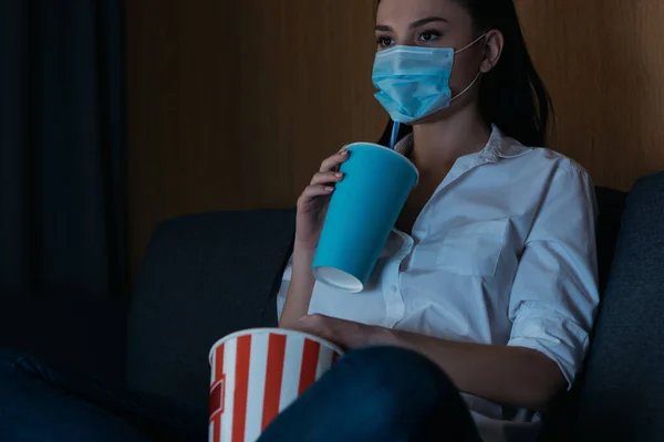 Junge Frau in medizinischer Maske hält Limo und Popcorn in der Hand, während sie zu Hause fernsieht — Stockfoto