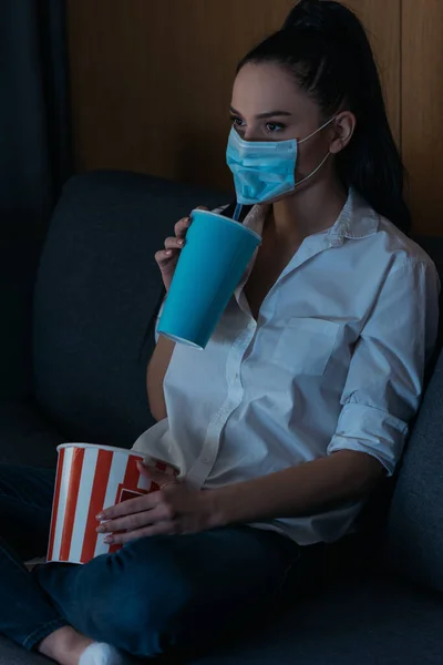 Jeune femme dans le masque médical tenant soda et pop-corn tout en regardant la télévision à la maison — Photo de stock