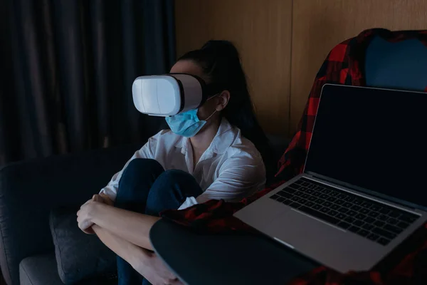 Mujer deprimida sentada en el sofá en auriculares vr cerca de la computadora portátil con pantalla en blanco en la silla - foto de stock