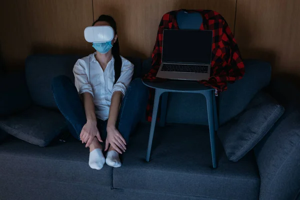 Femme déprimée assis sur le canapé et en utilisant un casque vr près d'un ordinateur portable avec écran vide sur la chaise — Photo de stock