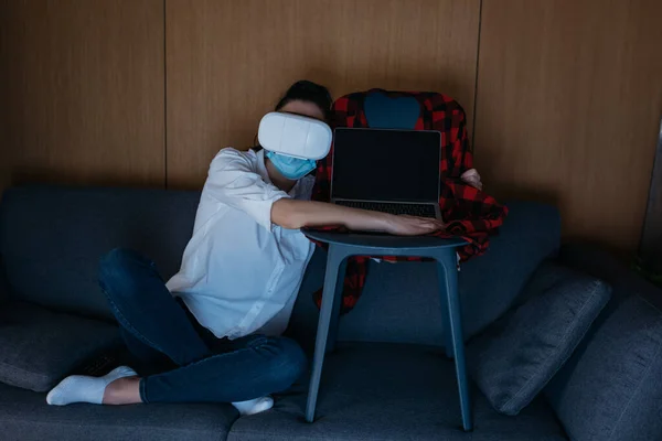 Mujer joven en auriculares vr sentado en el sofá y abrazando el ordenador portátil con pantalla en blanco en la silla - foto de stock