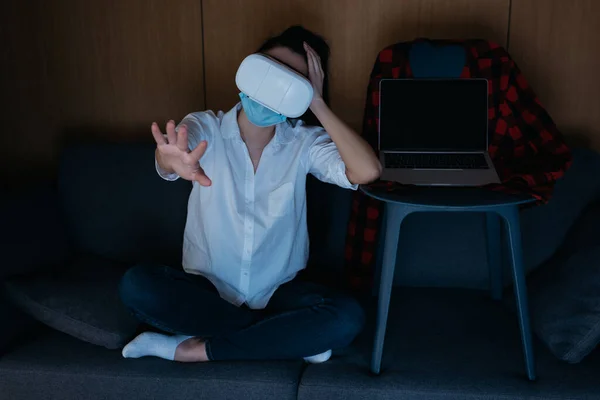 Jeune femme avec la main tendue en utilisant le casque vr tout en étant assis sur le canapé près de l'ordinateur portable avec écran blanc sur la chaise — Photo de stock