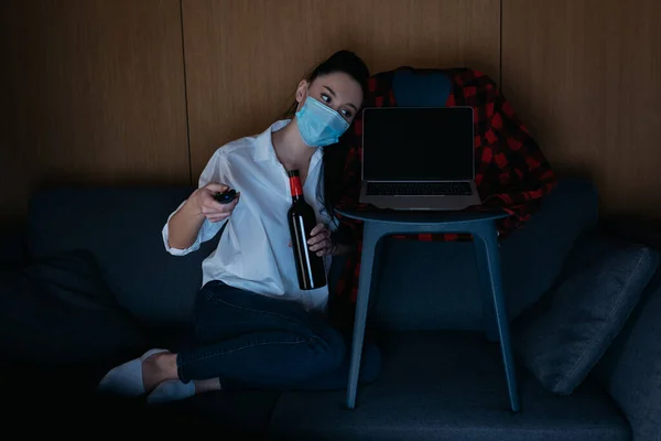 Mujer joven en máscara médica sosteniendo botella de vino y tv mando a distancia mientras está sentado en el sofá cerca de la computadora portátil con pantalla en blanco - foto de stock