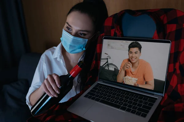 Jovem mulher no médico máscara segurando garrafa de vinho enquanto sentado no sofá perto laptop com sorrindo asiático namorado na tela — Fotografia de Stock