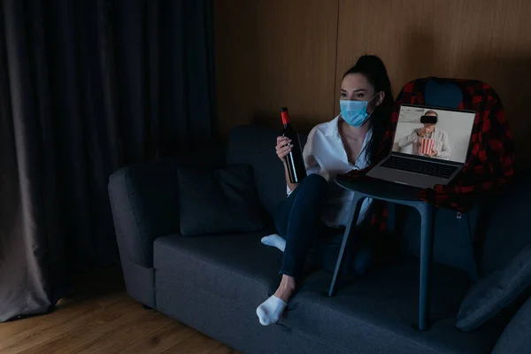 Jeune femme dans le masque médical tenant bouteille de vin tout en étant assis sur le canapé près d'un ordinateur portable avec petit ami dans un casque vr à l'écran — Photo de stock