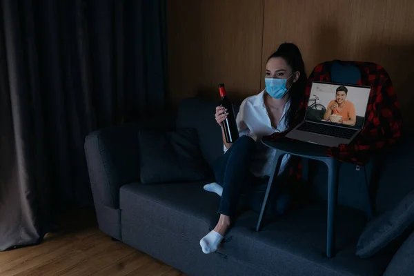 Joven mujer en médico máscara celebración botella de vino mientras está sentado en sofá cerca de portátil con sonriente asiático novio en pantalla - foto de stock