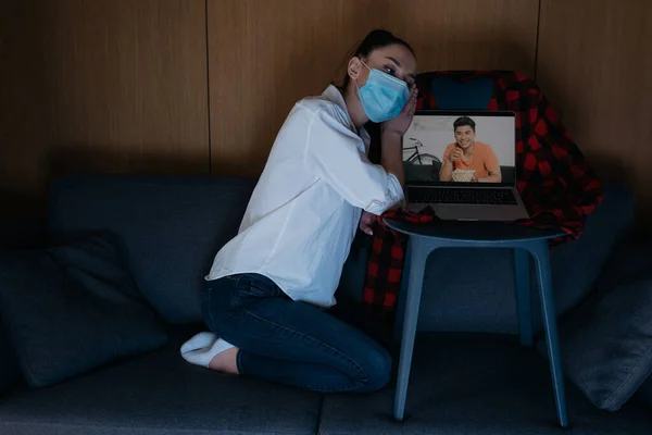 Грустная женщина в медицинской маске сидит на диване рядом с ноутбуком с улыбающимся азиатским парнем на экране — стоковое фото