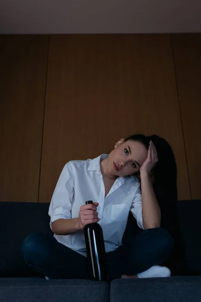 Femme déprimée toucher la tête et regarder la caméra tout en étant assis sur le canapé avec une bouteille de vin — Photo de stock
