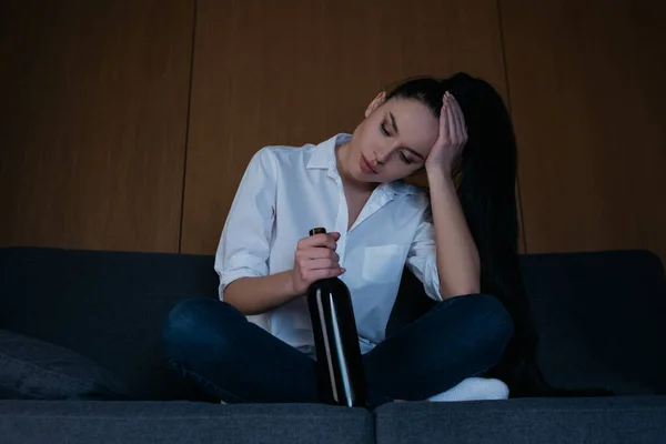 Depressed woman touching head while sitting on sofa with bottle of wine — Stock Photo