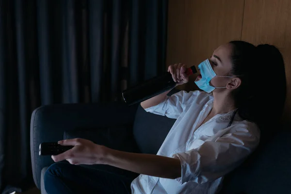 Depressed woman in medical mask with hole drinking wine from bottle while using tv remote controller — Stock Photo