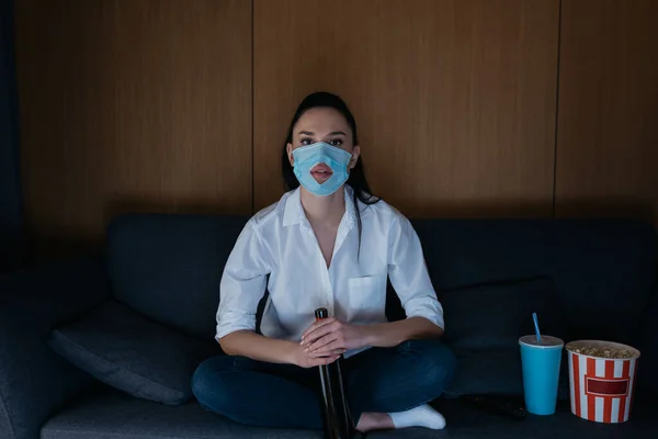 Jeune femme dans un masque médical avec trou tenant bouteille de vin tout en regardant la télévision sur le canapé près de soude et pop-corn — Photo de stock