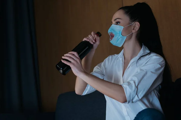 Femme déprimée dans un masque médical avec trou tenant bouteille de vin tout en étant assis à la maison — Photo de stock