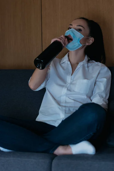 Depressed woman in medical mask with hole drinking wine from bottle — Stock Photo