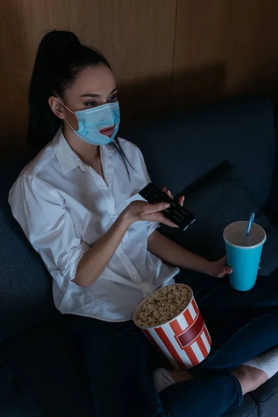 Vue grand angle de la jeune femme dans le masque médical avec trou en utilisant la télécommande tv tout en étant assis sur le canapé avec du pop-corn et de la soude — Photo de stock