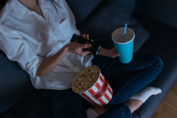 Cropped view of woman using tv remote controlled while sitting on sofa with popcorn and soda — Stock Photo