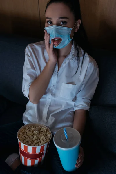 Worried woman in medical mask with hole touching face and looking at camera while watching tv with soda and popcorn at home — Stock Photo