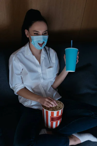 Улыбающаяся женщина в медицинской маске, смотрящая телевизор с газировкой и попкорном дома — стоковое фото
