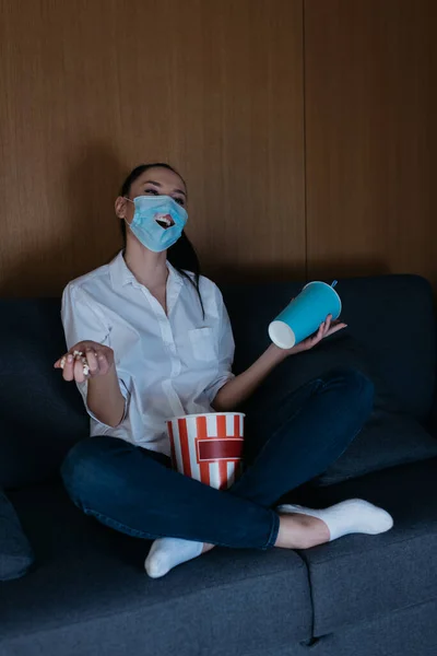 Sorridente giovane donna in maschera medica con buco guardando la tv con soda e popcorn a casa — Foto stock