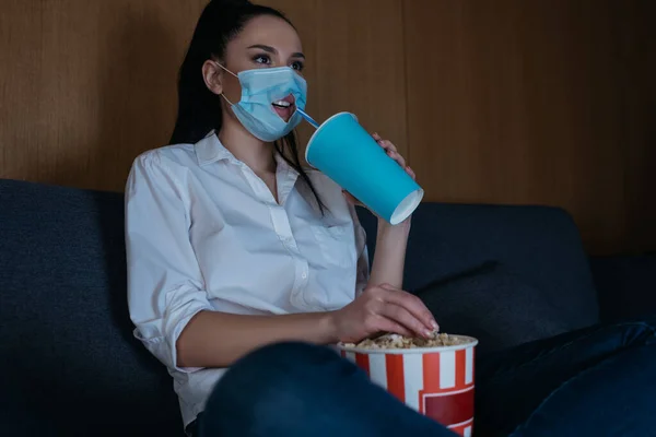 Jovem mulher em máscara médica com buraco sentado no sofá com pernas cruzadas, beber refrigerante, comer pipocas e assistir tv — Fotografia de Stock